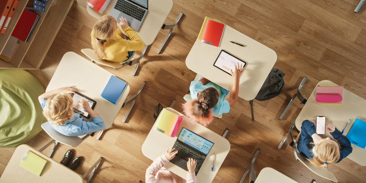 recursos-pedagogicos-iStock-1254051142-no-texto Vista superior da sala de aula no ensino fundamental: crianças sentadas na mesa da escola usando computadores pessoais e tablets digitais para tarefas.